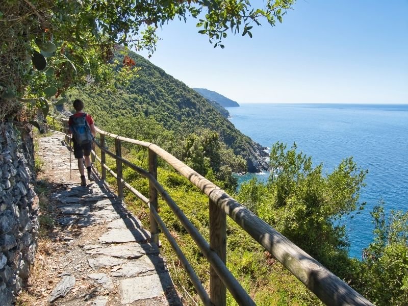 a man in shorts with walking sticks hiking along the path on the edge of the hillside near the ligurian sea in the cinque terre part of italy