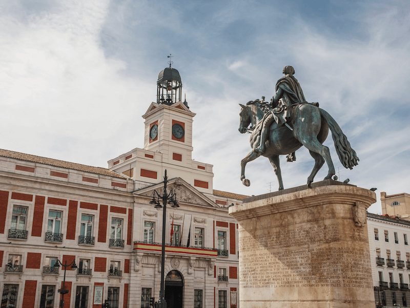 man on a horse statue in front of a red building with a visible clock tower part of the puerta del sol landscape