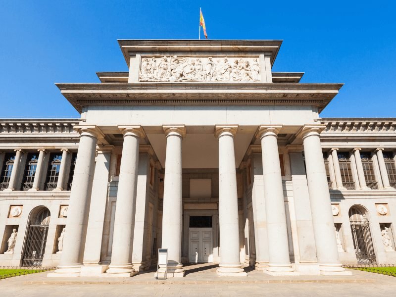 six pillars and fancy relief sculpture at the head of the entrance of the prado museum with a spanish flag flying atop it