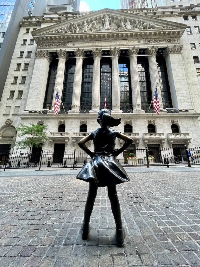 statue of the 'fearless girl' looking up at the new york stock exchange with colonnades, pillars, and two american flags