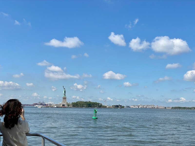 woman taking a photo of the statue of liberty while on the ferry in new york harbor on a sunny day with few clouds in the sky