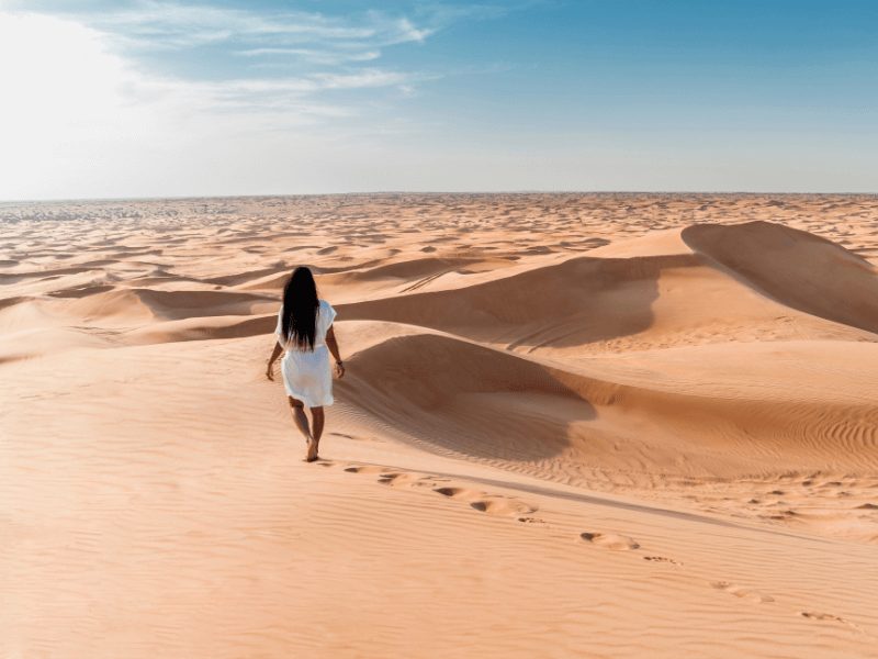 a woman with long black hair and brown skin walking away from the camera in a white knee-length dress, walking towards the dunes, leaving footprints behind her.