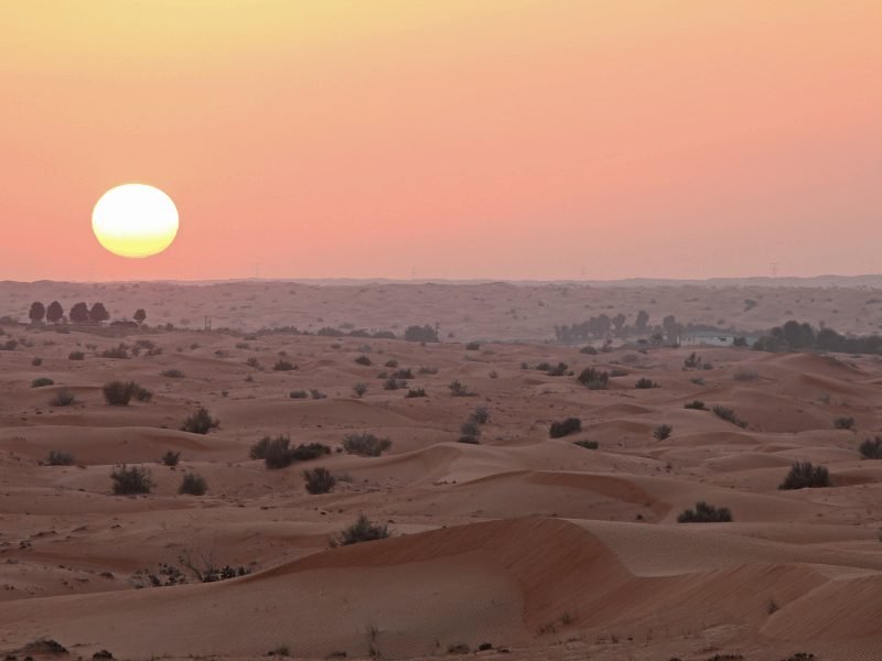 the sun setting over the Dubai desert, making the sky turn orange and yellow, and the red dunes turn darker