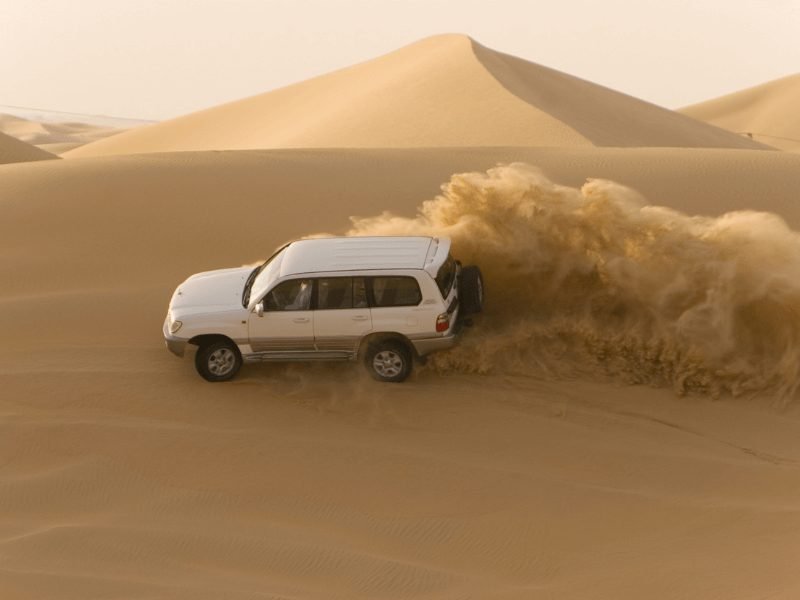 white all wheel drive car going through the sand dunes causing sand to kick up wildly