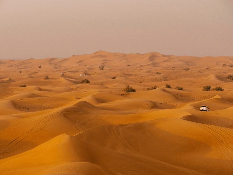 several white all wheel drive cars going through the dunes of the dubai desert in the late afternoon with a dusty sky