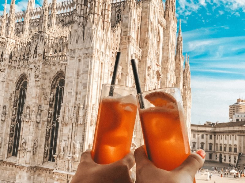 two glasses of cocktails cheersing in front of the milan duomo