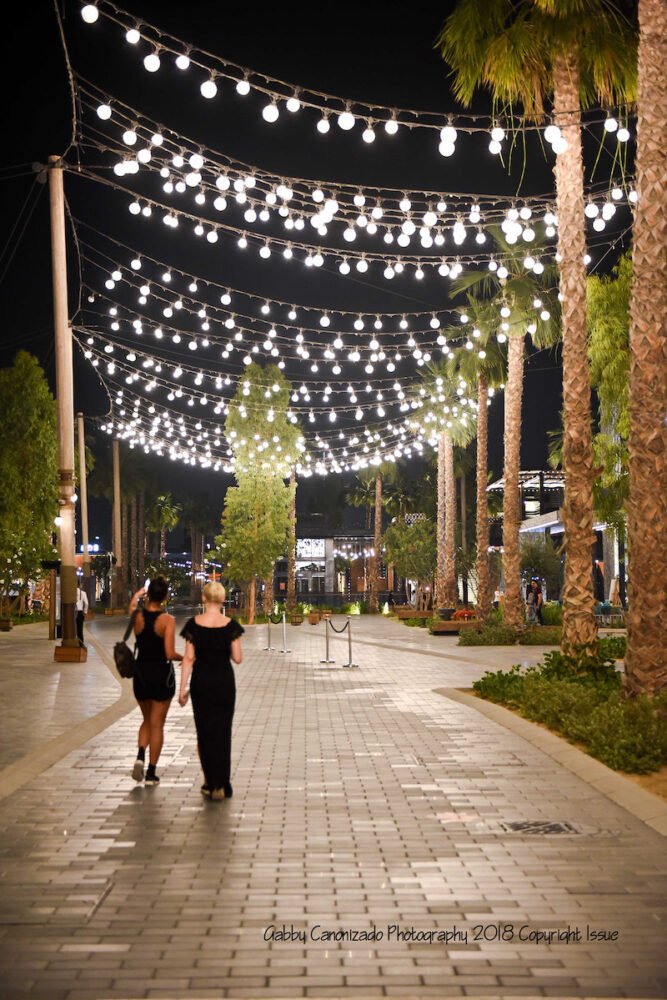 two women walking around la mer beach area at night with lights and palm trees
