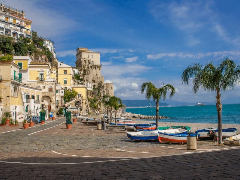 small fishing boats on the beach, a few small palm trees, yellow and brown buildings on the edge of the sea in a cute amalfi coast town