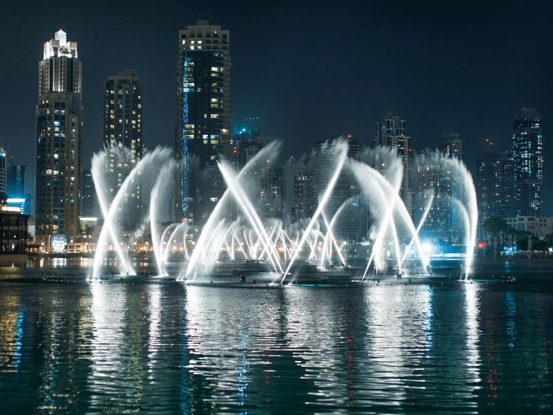 the dubai fountain as seen at night, lit up and with the skyline behind it