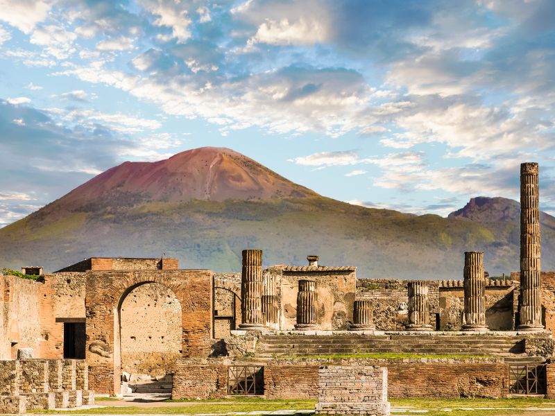 The ruins of Pompeii (arches, pillars, walls) that have been excavated, with Mt. Vesuvius -- the volcano that erupted and destroyed it -- in the backgroun.