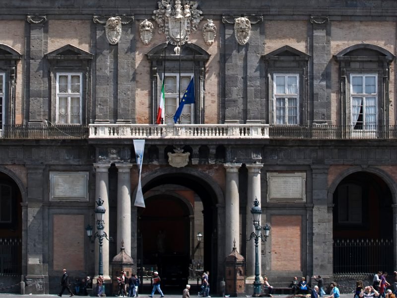 the exterior of naples royal palace with italian flag and eu flag and people walking in front