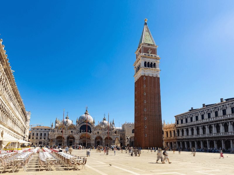 venice belltower and church in san marco square with chairs out for dining and people walking around