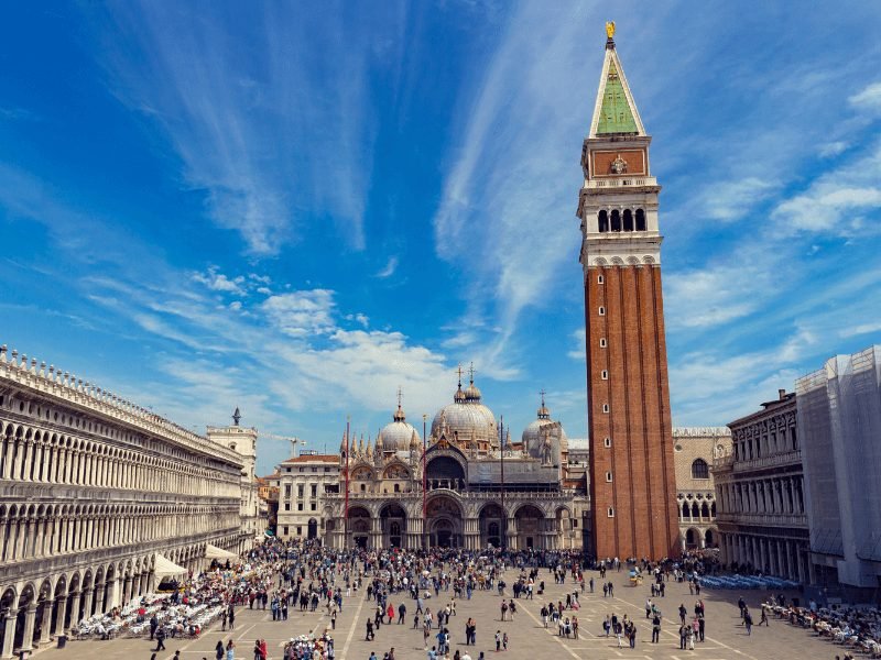 The busy square of Piazza San Marco with lots of tourists milling about in the square area on a sunny day. The campanile (bell tower) stands high over the top of the piazza)