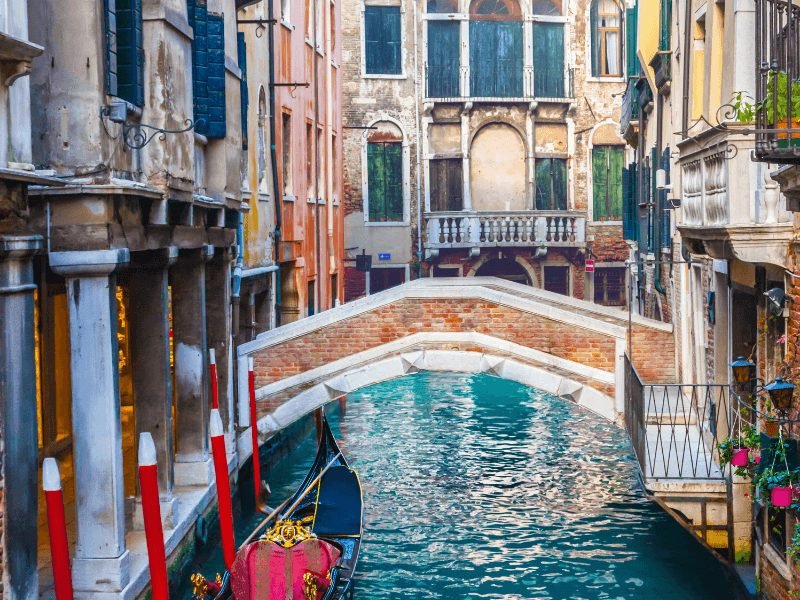 Gondola sitting in the canal, wiht a brick bridge, and other storefronts and buildings in Venice Italy