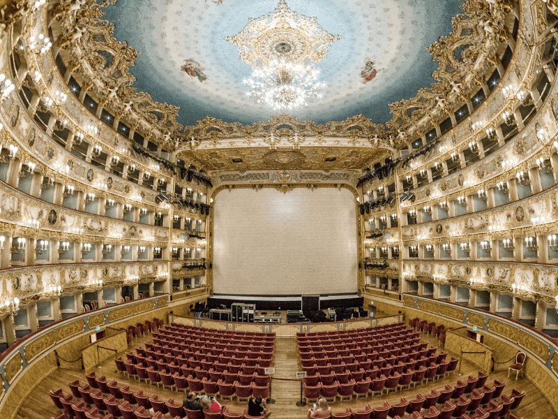 The interior of the La Fenice opera house in Venice, with ornate ceiling painting, boxes, and red plush floor seating.