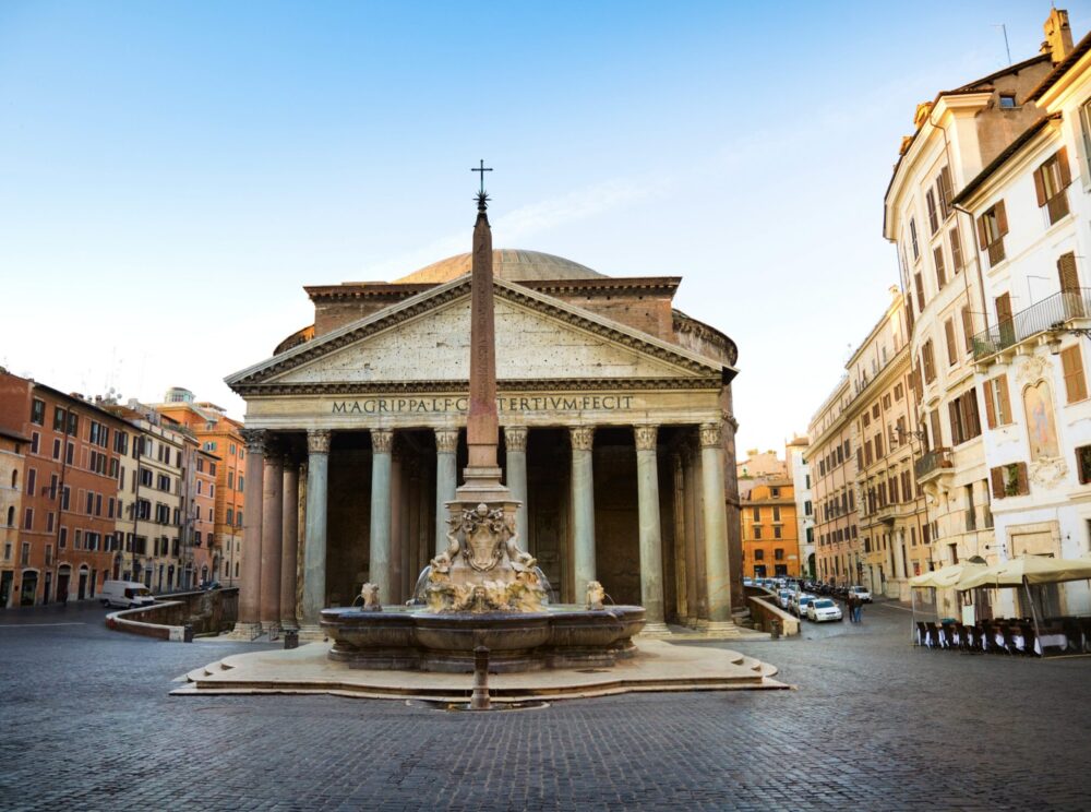 fountain with cross on the top in front of pantheon in rome with seven columns and roman lettering on front of temple