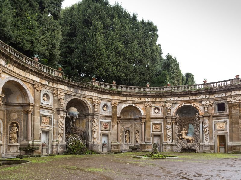 villa in the town of frascati with statues and archways and artwork with trees behind it