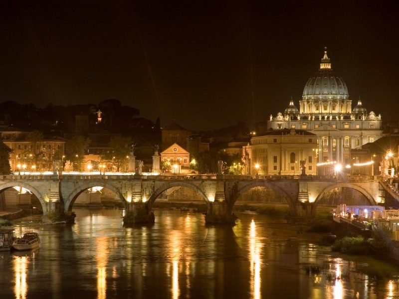 lit up bridge over the tiber river in rome at night with the vatican buildings behind it