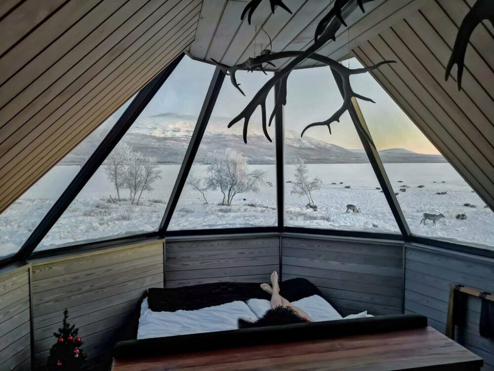 A woman's legs relaxing in a bed as she enjoys the view out her Finland glass igloo, looking out onto a reindeer farm and a snowy landscape with sunrise light with pastel colors on the horizon. She is in bed and you can see an antler decor chandelier above the bed.