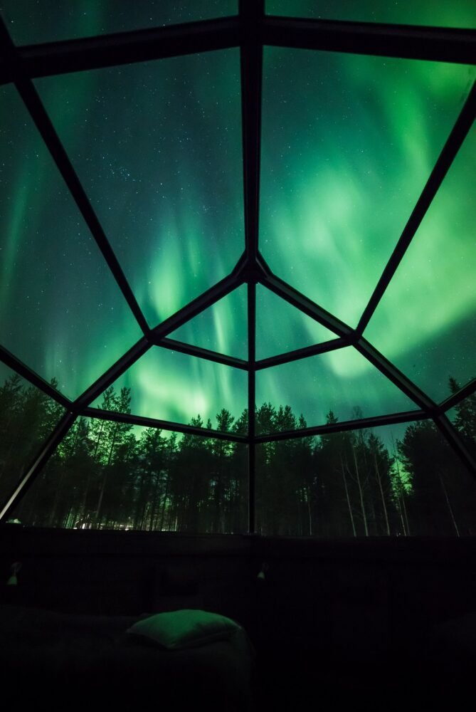 View of intense aurora colors as seen from the interior of a glass igloo cabin in Finland during the winter season. Brilliant blueish green color aurora in the dark night sky.