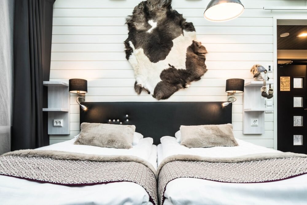 The interior of a glass igloo with beautiful linens, reindeer pelt detailing, fluffy pillows and white wood paneled walls. The design is very Nordic and minimalist yet cozy.