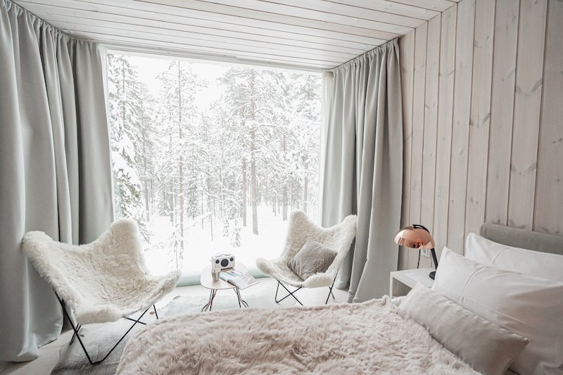 Beautiful white and wood room with fluffy fur-covered duvet, lots of pillows on bed and two fluffy chairs with a vintage radio on the table, view looking out onto a big glass window with snow-covered trees.