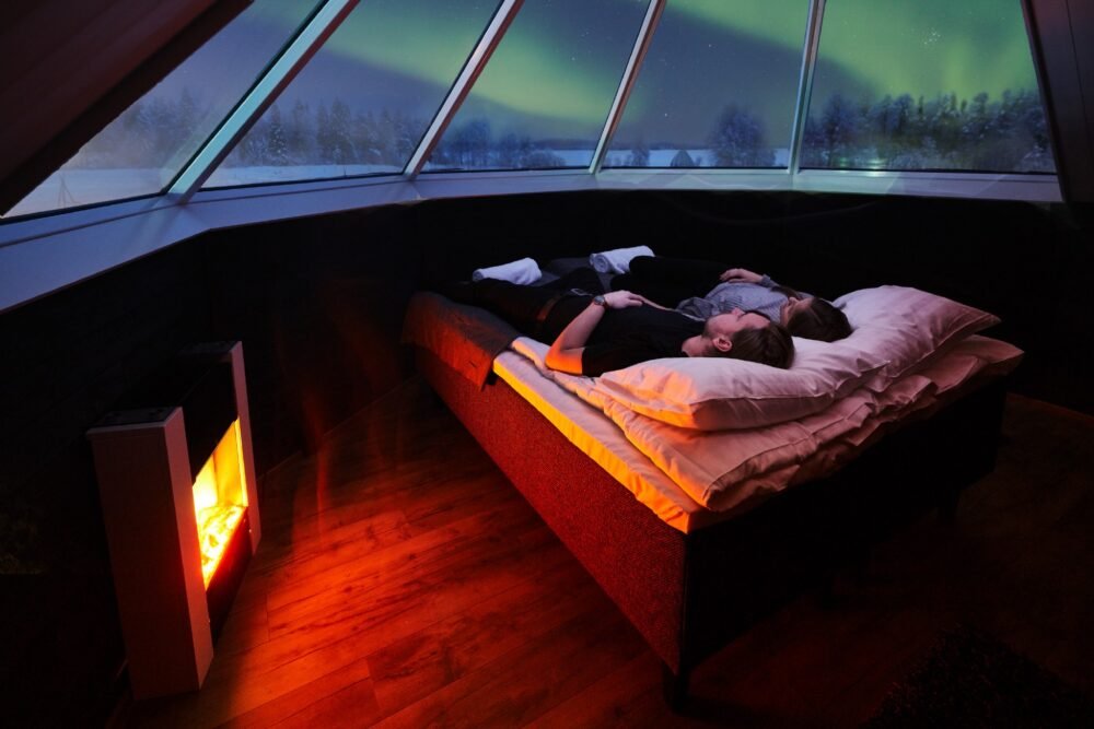 The interior of an aurora cabin in Apukka Resort with a couple next to a fake fireplace for warmth looks up at the green streaks of the Northern lights in the sky.