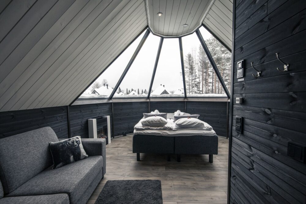 Interior of an aurora cabin at Apukka Resort with bed, electric fireplace, sofa, all during the daytime so you can see the minimalist furnishing and detail. White and gray furnishings make it look very elegant.