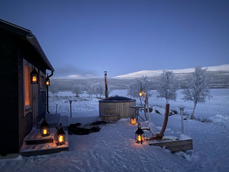 The night sky is dark but there are warmly lit lanterns on the snowy ground leading you to a hot tub outside in the snow.