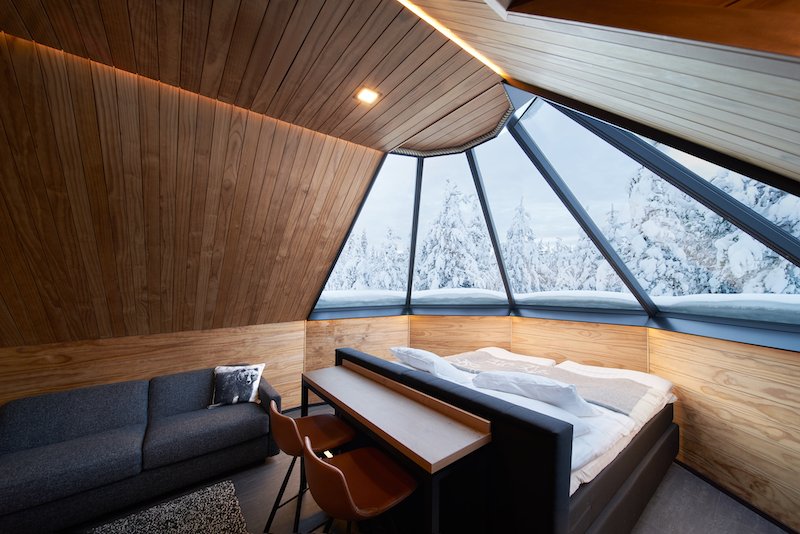 The interior of a glass igloo in Levi. There is a double bed facing a panoramic glass window, as well as a seating area and two chairs at a small bar against the bed. The ceiling and walls are made of wood and glass and the design is simple. The glass windows show a winter landscape with snow during the daytime.