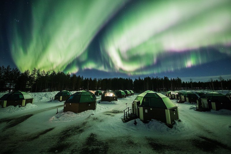 Green spirals of the Northern lights in the night sky above many glass igloos, in a landscape covered in snow, on a winter day in Finnish Lapland.