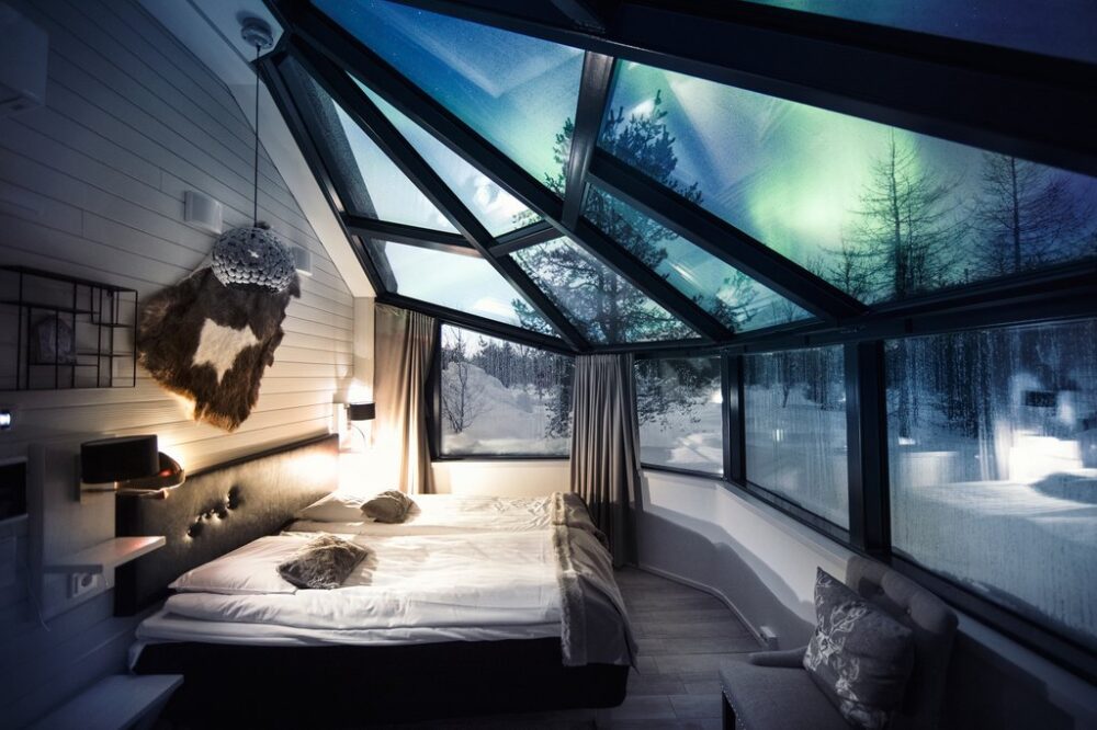 The aurora outside the window of a Finland glass igloo is vibrant with green color. Inside, you can see the white bed, Finnish design detail, and glass panes of window that show you the aurora from your window which has 180 degree views of the colorful night sky.