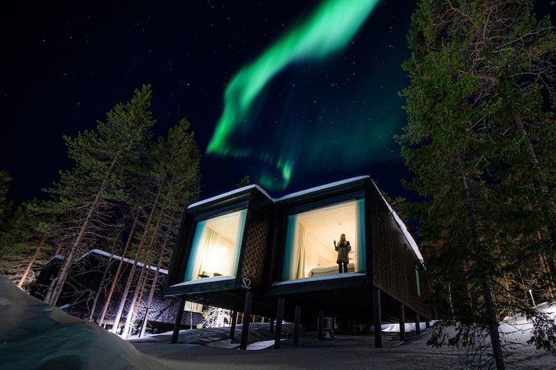 A woman standing in the window of one of the treehouse glass aurora cabins with the aurora dancing overhead while in Finnish Lapland in winter