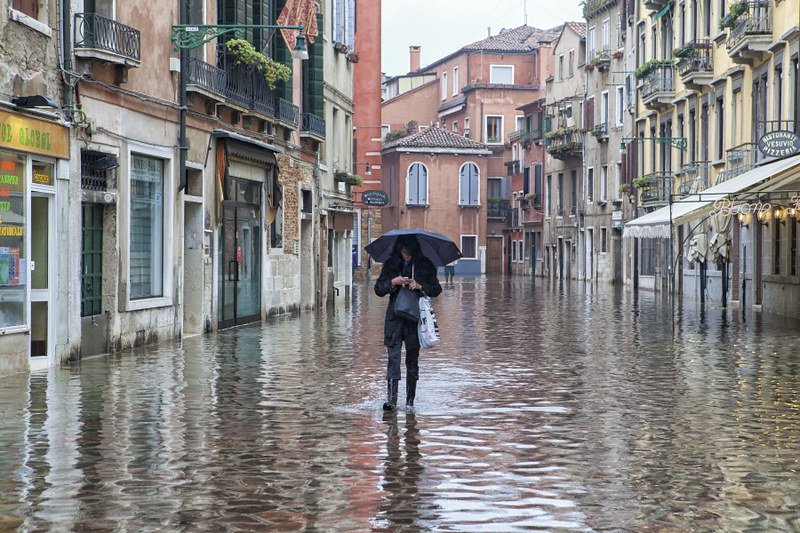 Woman in a flooded area of Venice wearing jeans, a jacket, rain boots and holding an umbrella. The city buildings behind her are visible and it is a gray and rainy day.