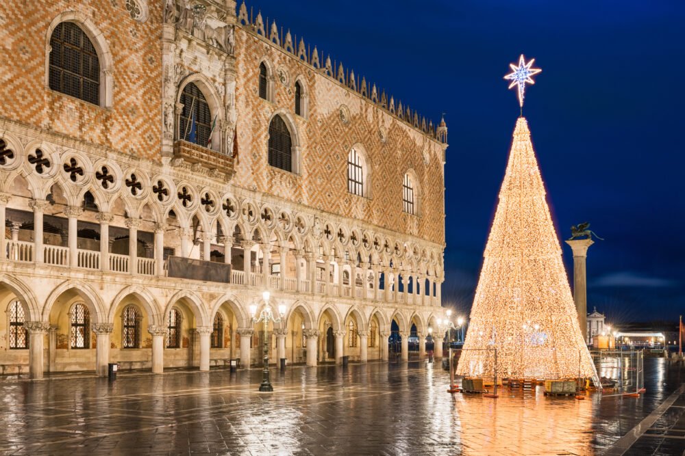 Christmas decorations at the San Marco square in Venice, Italy, with lit up tree, Doge's Palace pillars and archway, and dark blue-black sky.