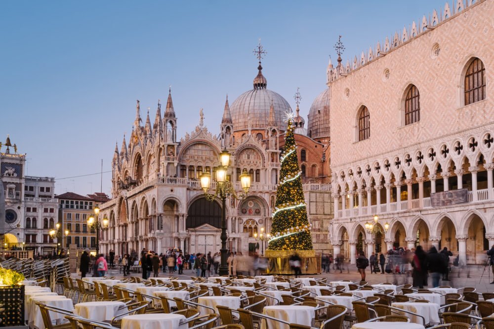 Christmas tree in Piazza San Marco with lots of tables at cafes out able to eat and have a drink and enjoy the Christmas views in the square, in soft blue light after sunset but not too dark. Church building and doge's palace windows and pillars visible.