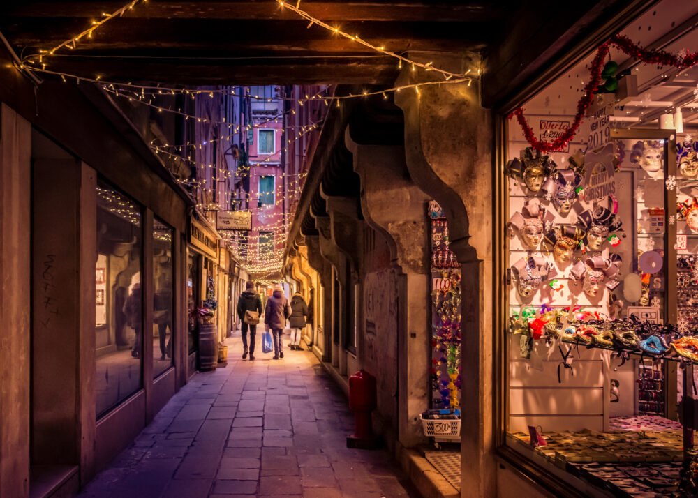People wandering down the streets of Venice which are lit up with Christmas lights giving everything a festive atmosphere, and a store is selling Venetian colorful carnival masks in the window.