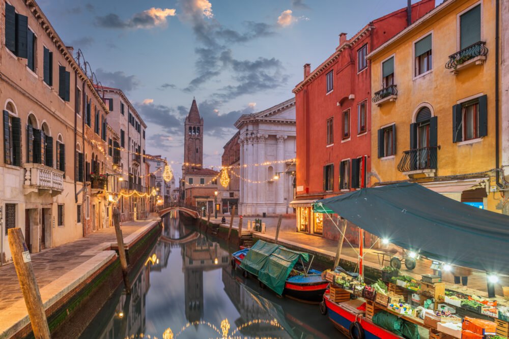 View of the canals of Venice with lights, campanile in the background, lit up gondola and colorful buildings, around sunset hour.