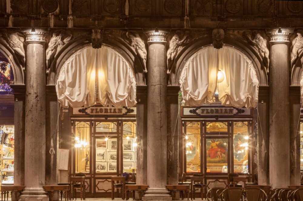 Old Traditional Bar Caffe Florian in St. Marks Square, with pillars, lit up curtains, and warm indoor lighting, inviting you to come inside for a drink.