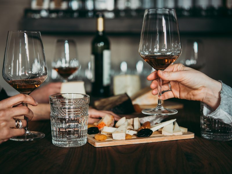 hands holding wine glasses with a plate of cheese and dried fruit behind the people