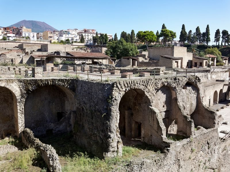 Herculaneum Boathouse facing the sea with the rest of the ancient city in ruins behind it and Mt Vesuvius, the volcano that destroyed it, nearby