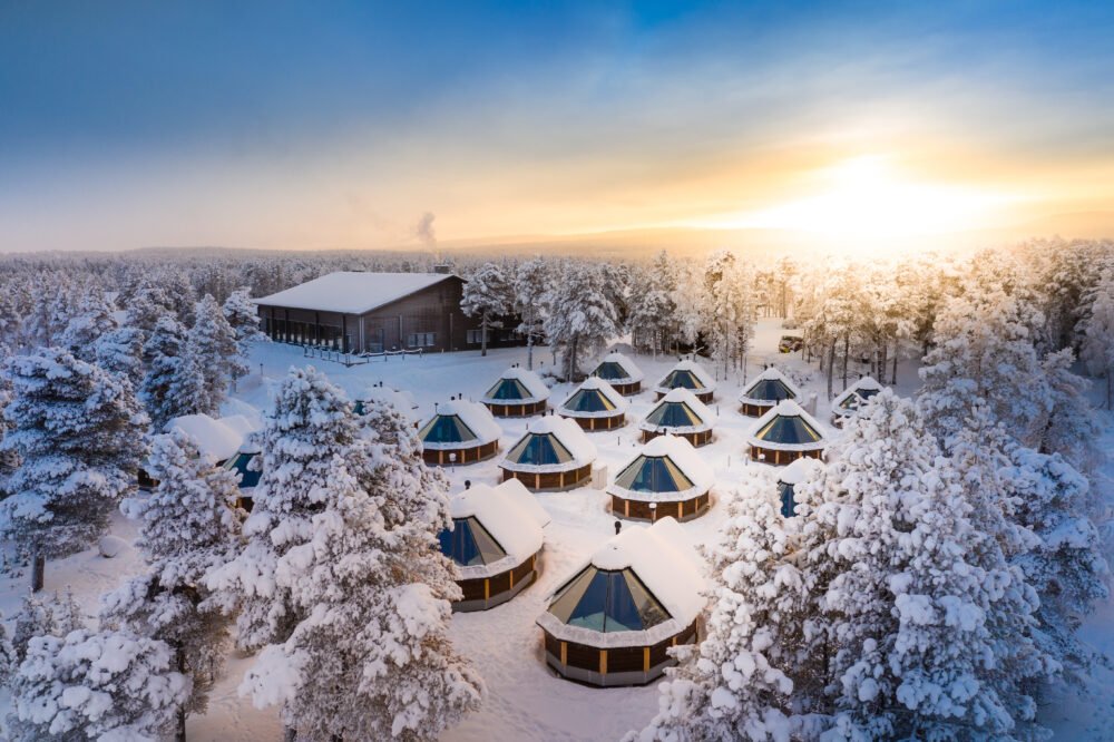 Drone shot of a glass igloo in Finland with glass ceiling, snow-covered cabins, sunrise looming on the horizon and snow-covered trees in Lapland.