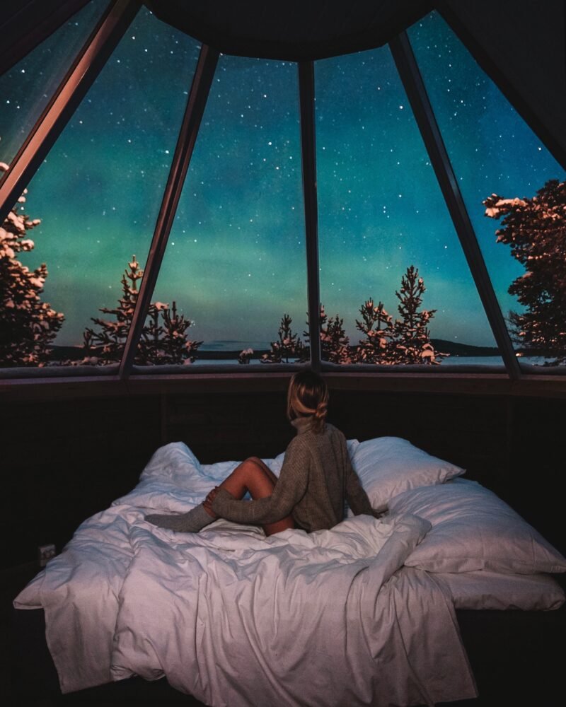 A woman sitting on a bed looking at the aurora and snow-covered trees outside of her glass igloo in Finland