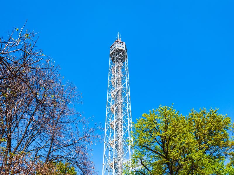 large steel-looking tower with a hexagon-shaped observation deck area on the top on a sunny day in Milan in a park with trees surrounding it.