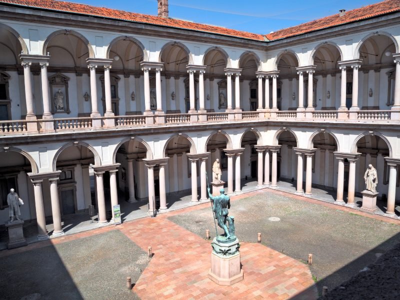 The interior courtyard of the Pinacoteca di Brera, with a patinated green statue of a man holding a spear, with white archways and porticoes around the courtyard of this Milan landmark.
