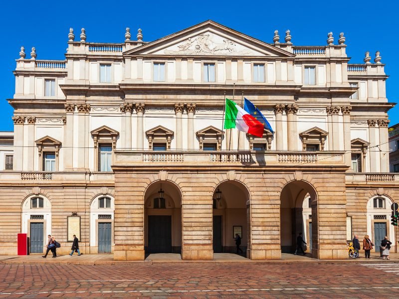 Three arches in front of the Milan Opera house, with an Italian flag in front, and beige building where the operas and ballets take place at this famous landmark in Milan, Italy.