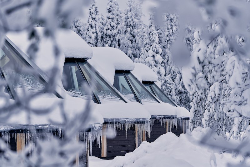 Snow-covered glass igloos in Finland in Levi, with a snowy landscape with trees coated in fluffy white snow, and icicles dripping from the aurora cabins.