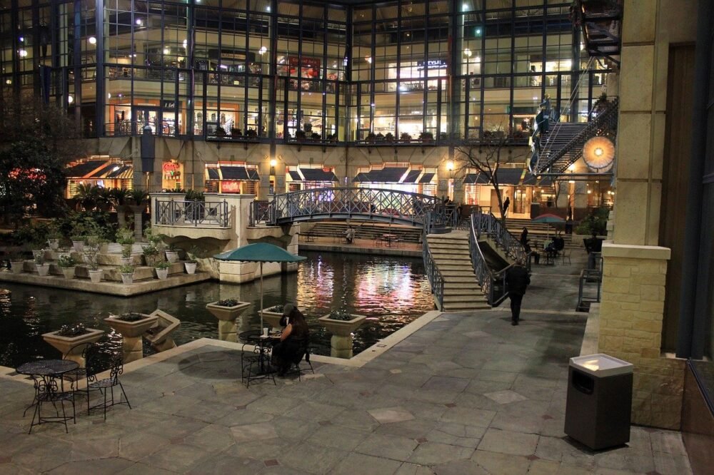 The San Antonio riverwalk at night, sidewalks paved alongside a river with bridges, shops, etc to visit