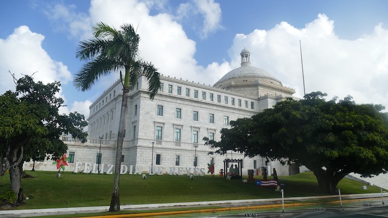 View of San Juan in December, with a sign that says "Feliz Dia de Reyes" which means "Happy day of the kings" in Spanish. There is a large fortress-like white structure in the background, as well as palm trees and other plant life, and a Puerto Rican flag icon, and a nativity scene.