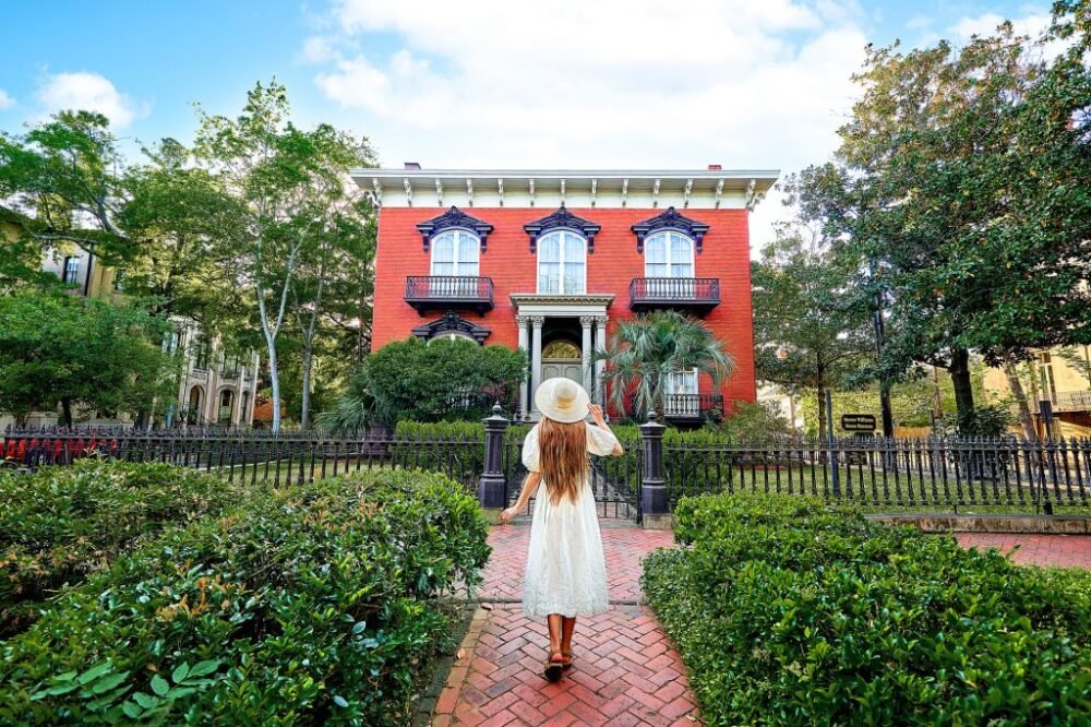 Victoria, the author of the section, with long hair and a white dress, walking in front of a red house called the Mercer House, with a garden in the front of the house on a sunny day in Savannah GA